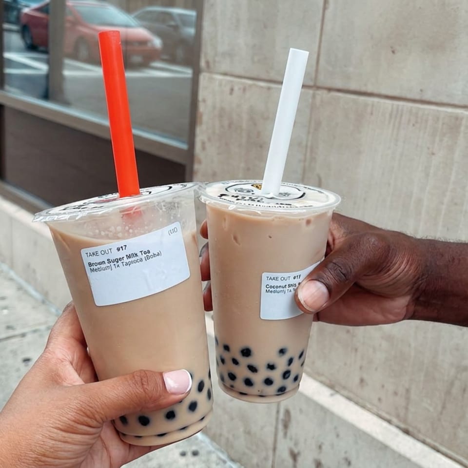Two hands hold iced brown sugar milk tea and coconut milk tea with black tapioca pearls outside a storefront.