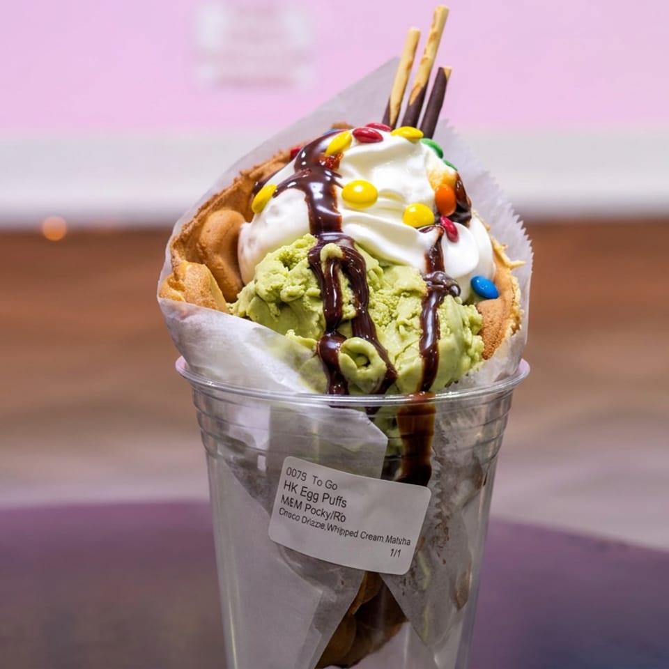 Matcha soft-serve parfait in a clear cup, topped with whipped cream, chocolate drizzle, colorful candies, and pretzel sticks.