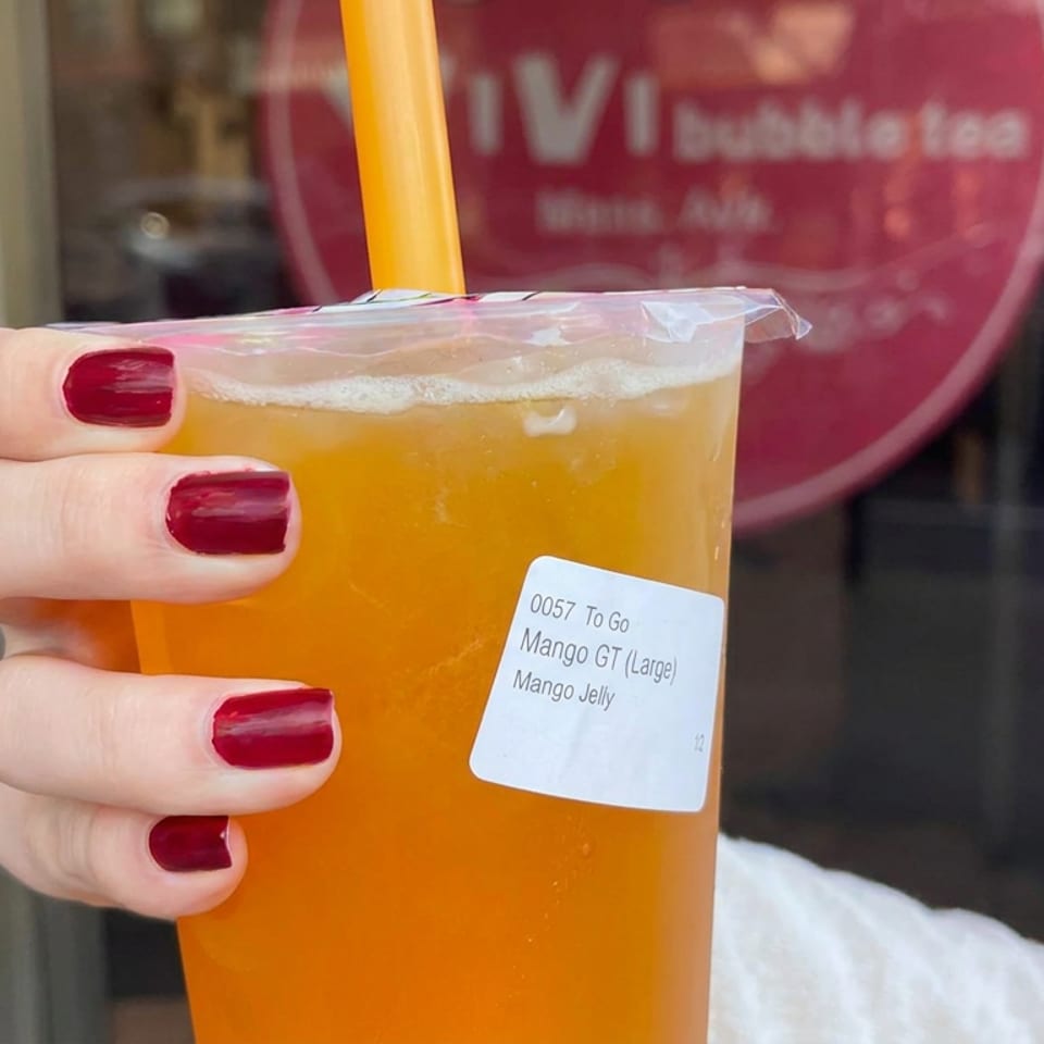 Hand with red-painted nails holding a large mango GT beverage labeled “Mango Jelly” outside a restaurant.
