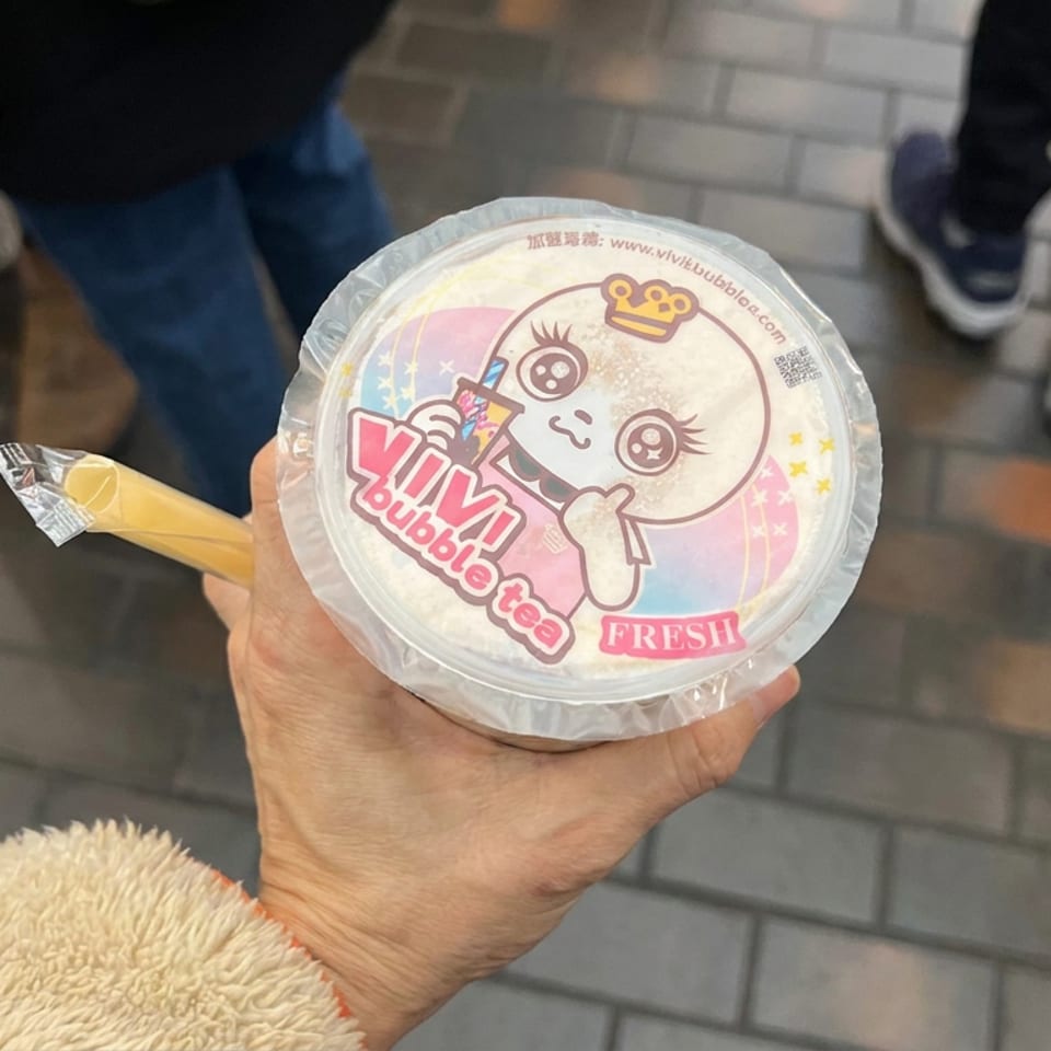 Hand holding a fresh YiYi bubble tea cup with a colorful cartoon lid, surrounded by people on a paved walkway.