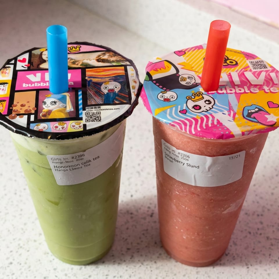 Two colorful boba drinks sit on a speckled counter, featuring green matcha milk tea and pink strawberry slush with cartoon-patterned sealed lids.