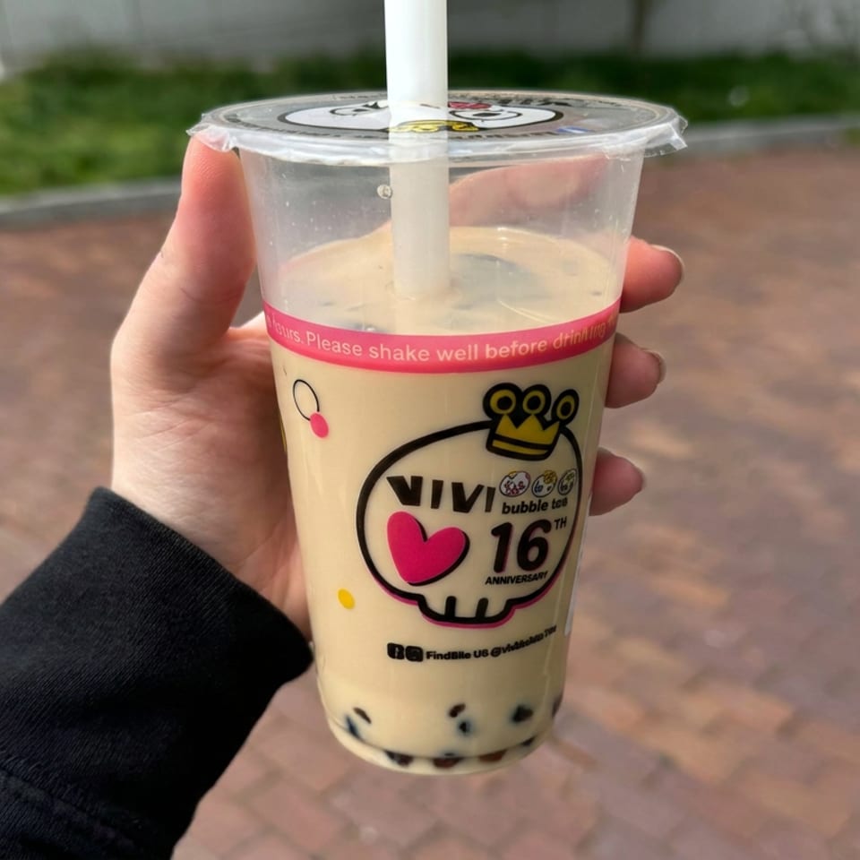 Hand holding a milk tea with tapioca pearls in a branded plastic cup outdoors on a brick walkway.