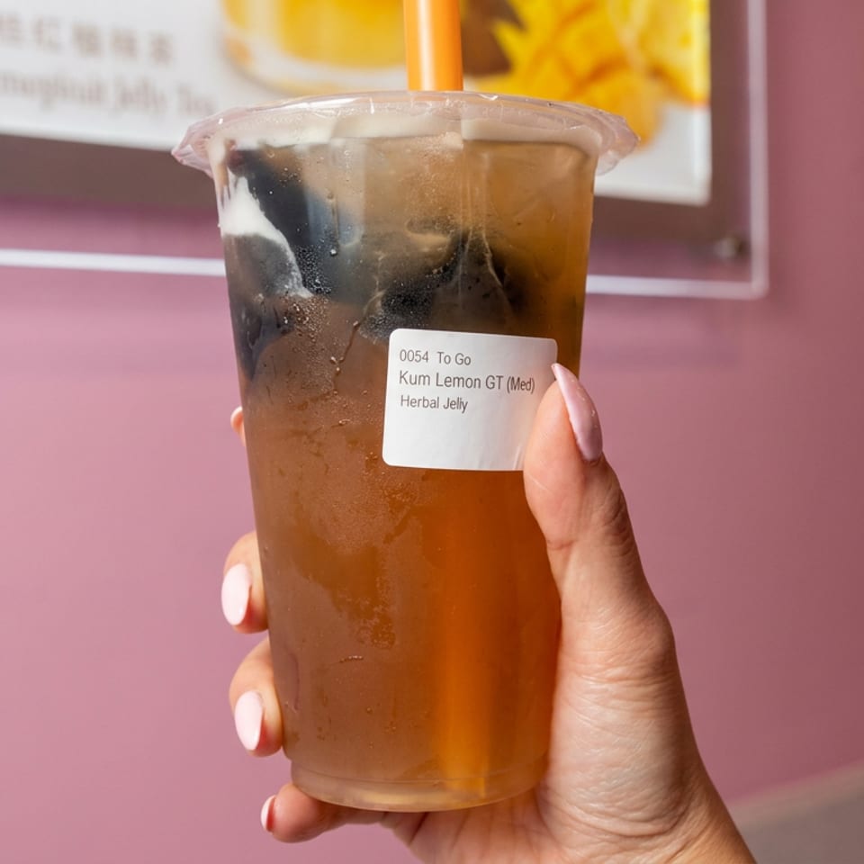 Hand holds a chilled cup of Kum Lemon GT with herbal jelly, condensation, ice, and an orange straw against a pink wall.