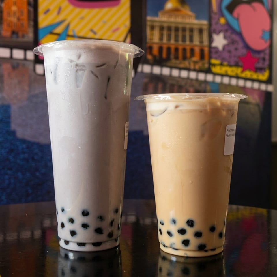 Two iced bubble tea drinks with tapioca pearls sit on a glossy table, with colorful artwork and café seating in the background.