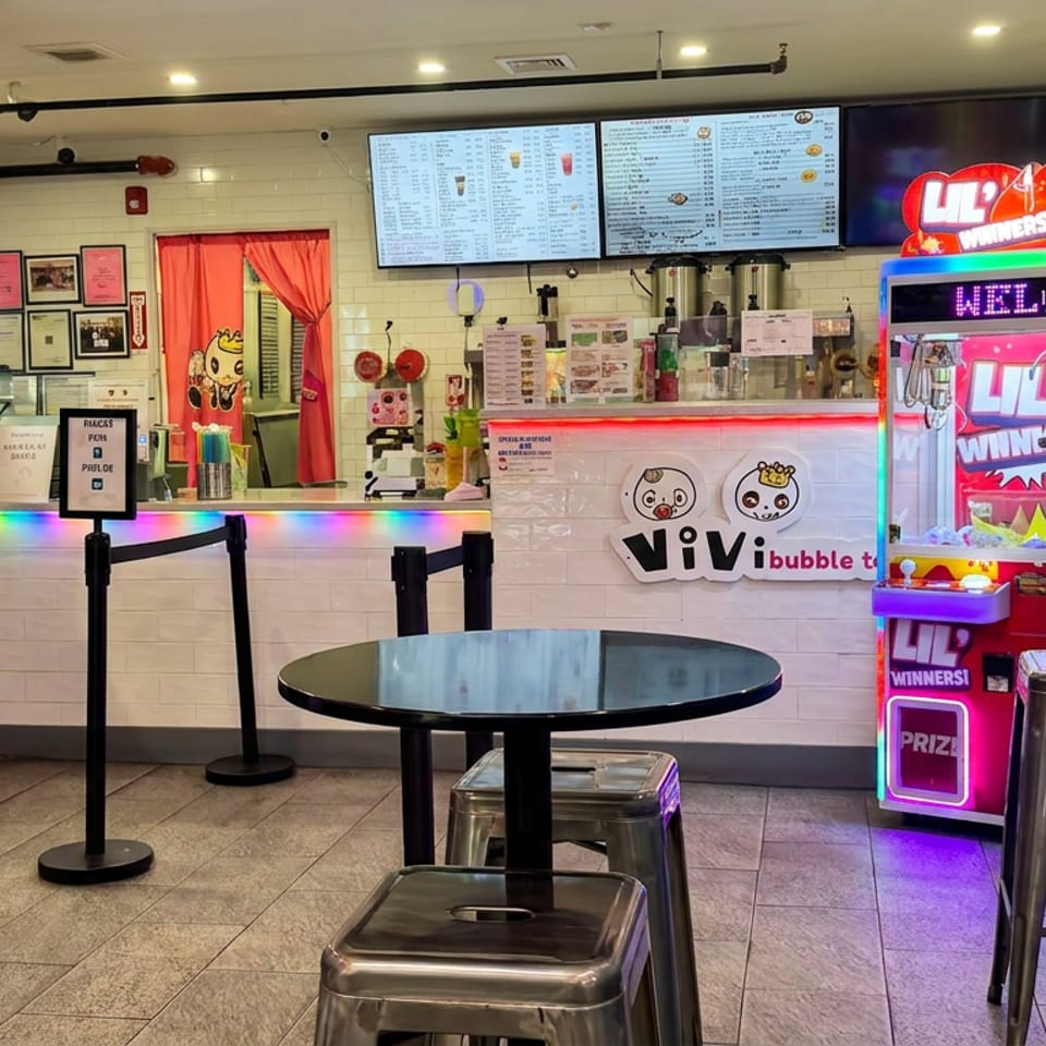 Colorful ViVi bubble tea shop interior with illuminated counter, digital menus, claw machine, and metal tables and stools.