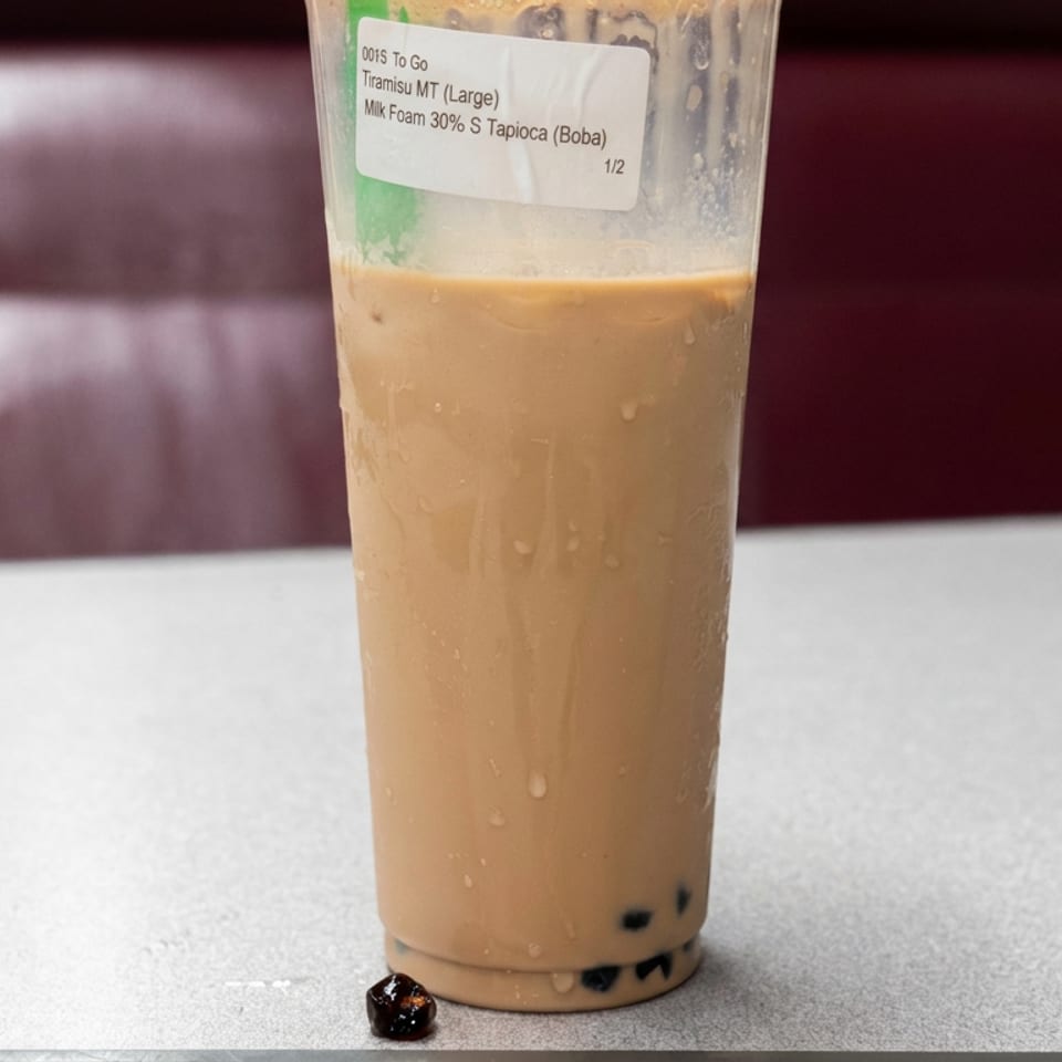 Large iced tiramisu milk tea with tapioca pearls in a clear cup, standing on a restaurant table.