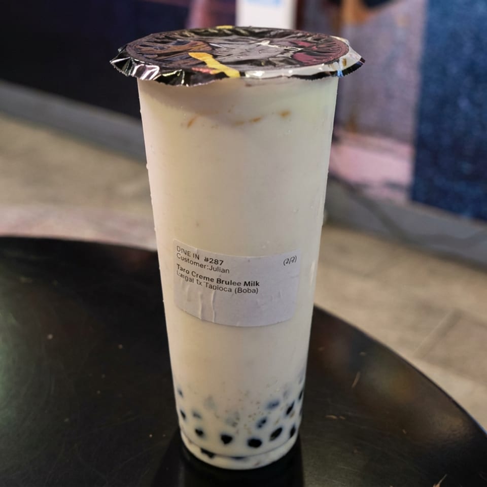 A sealed cup of taro crème brûlée milk tea with tapioca pearls sits on a black tabletop.