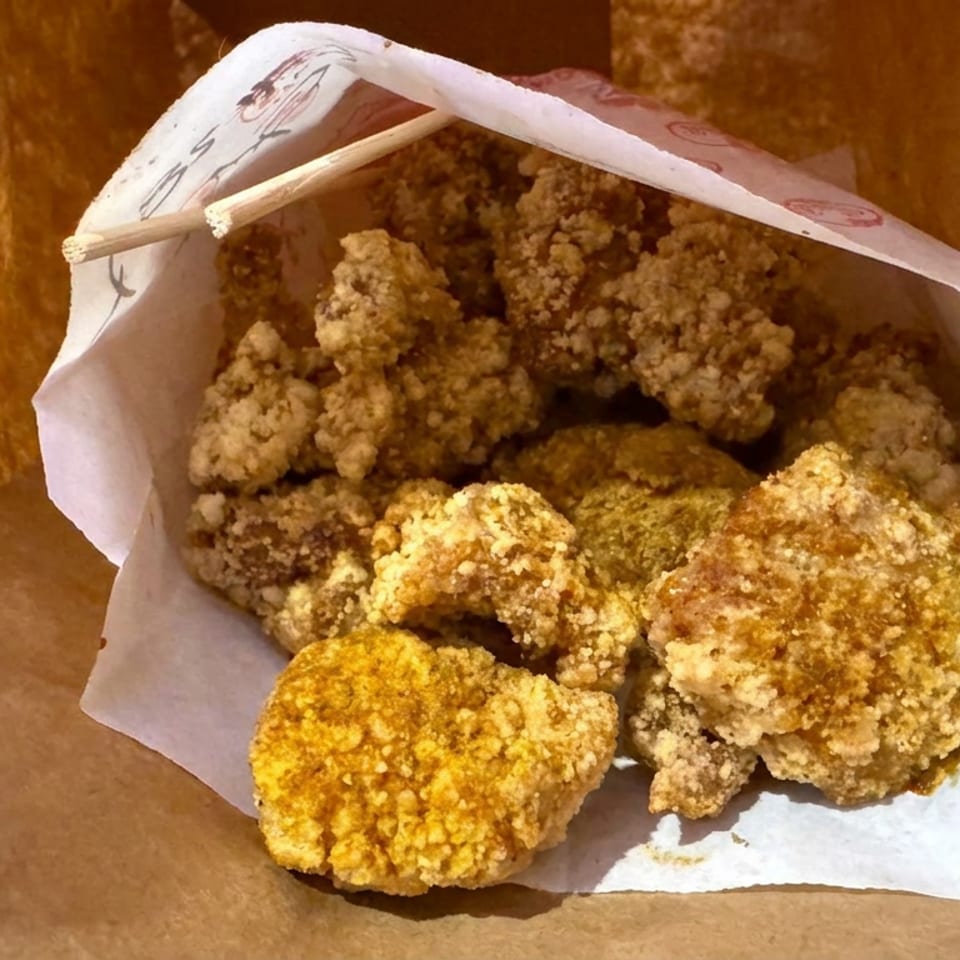 A paper-lined bag holds golden-brown crispy fried chicken pieces with wooden chopsticks resting across the top.